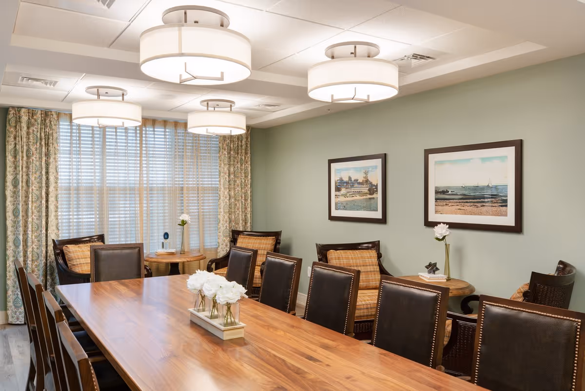 A well-lit dining room with a long wooden table surrounded by dark leather chairs. The room features three large round ceiling lights, patterned curtains covering a window, and two framed pictures on a light green wall. There are small round tables with cushioned chairs and decorative flowers placed on the tables.