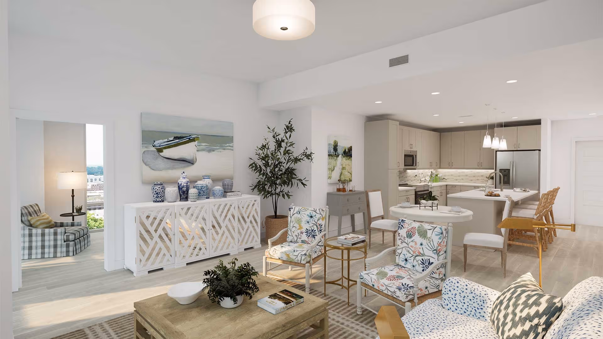 Bright and modern open-concept living area with floral patterned chairs, a wooden coffee table with plants and books, a white decorative cabinet with blue and white vases, and a kitchen with beige cabinets, a large island with bar stools, and pendant lighting.