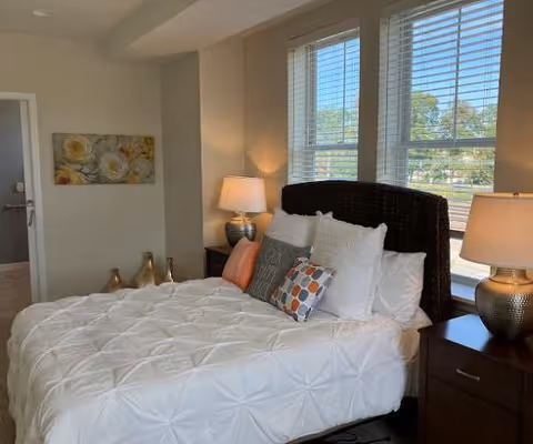 A cozy bedroom with a large bed featuring a dark woven headboard, white comforter, and multiple decorative pillows in white, gray, orange, and patterned colors. Two bedside tables with matching lamps flank the bed. A window with blinds lets in natural light, and a floral painting hangs on the wall near the door.