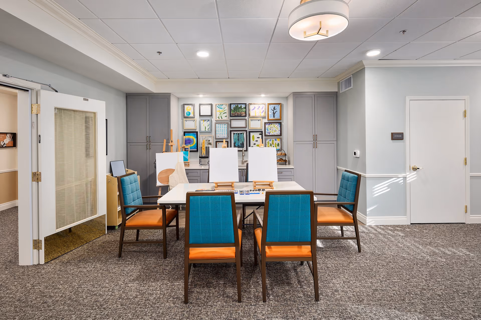 Bright craft/activity room with a central table surrounded by turquoise-and-orange chairs, easels with canvases, built-in cabinets and framed artwork.