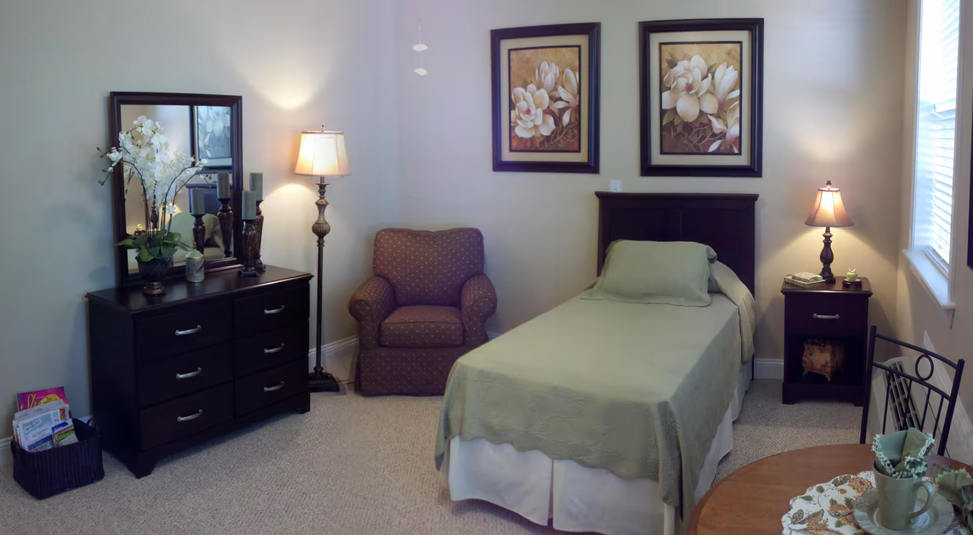 Furnished single-occupancy bedroom with a twin bed, dresser, armchair, and lamps under framed floral art.