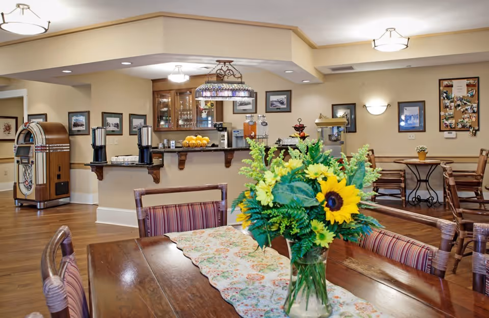 A cozy dining area in a senior living facility with wooden tables and chairs, a vase with a bouquet of sunflowers and other yellow flowers on the table in the foreground, a counter with beverage dispensers and a bowl of oranges in the background, framed pictures on the walls, and a vintage jukebox on the left side.