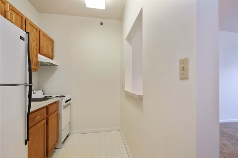 Small kitchen area with white refrigerator, white stove, wooden cabinets, and a pass-through window opening to an adjacent room with carpeted floor.