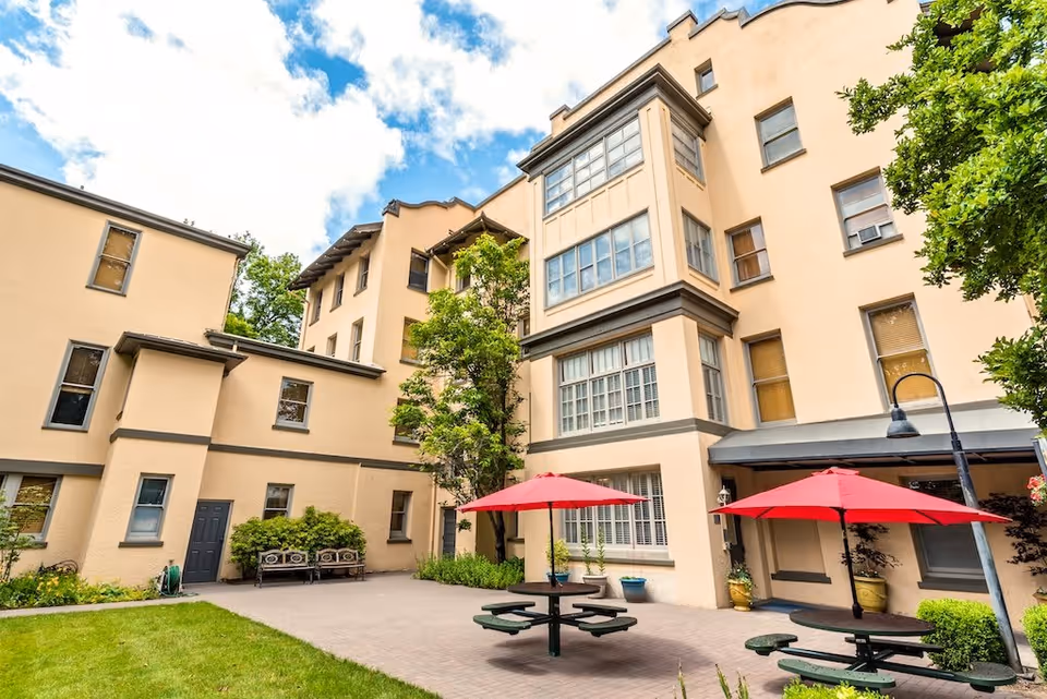 Outdoor courtyard area of St. Andrews Memory Care facility featuring beige multi-story building walls, green grass, two round picnic tables with attached benches and red umbrellas, a few potted plants, benches along the wall, and a partly cloudy blue sky.