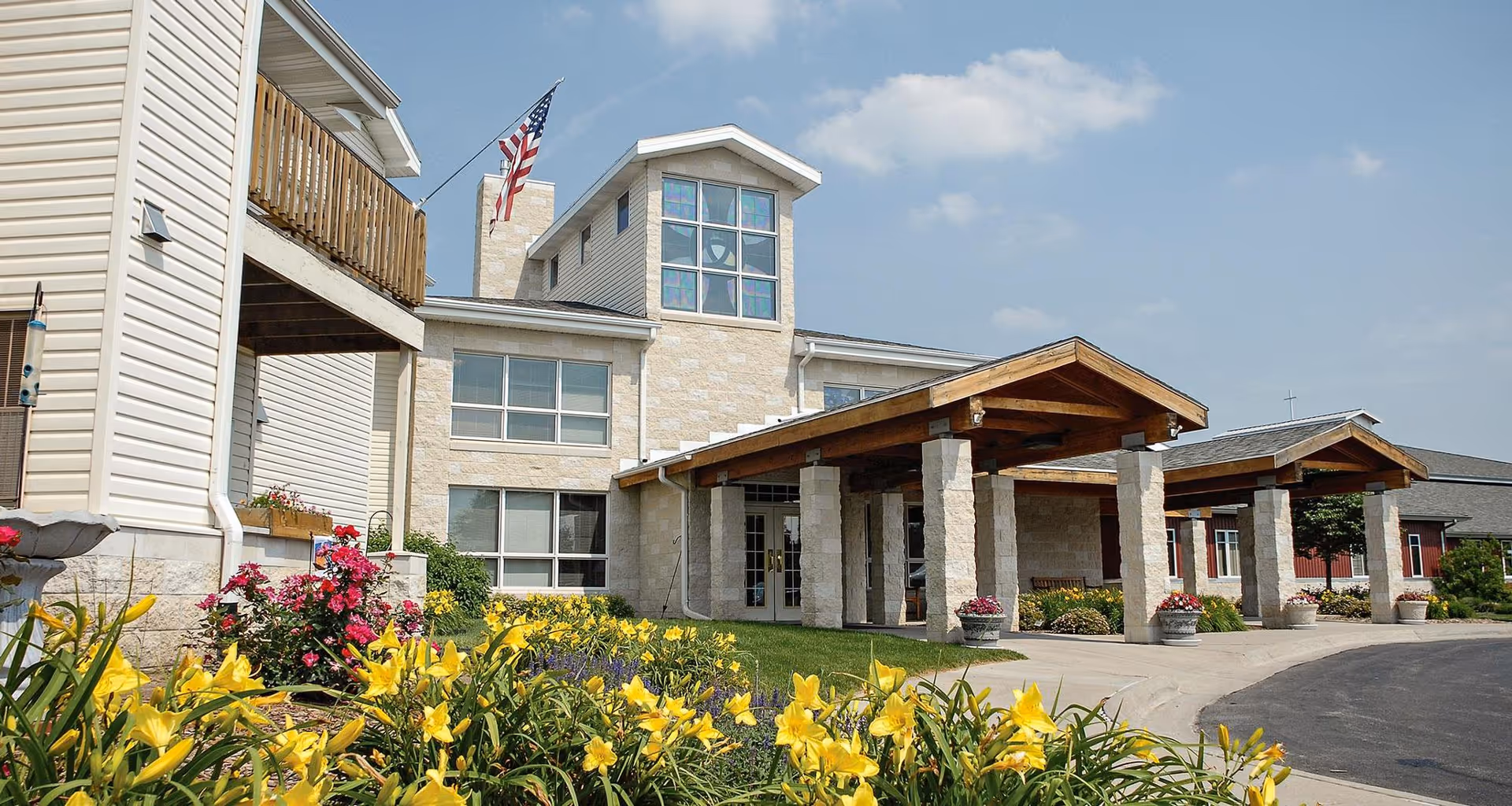 Exterior view of a senior living facility building with a covered entrance supported by stone pillars. The building has large windows and an American flag flying on a pole. In the foreground, there are yellow flowers and other landscaping plants under a partly cloudy sky.