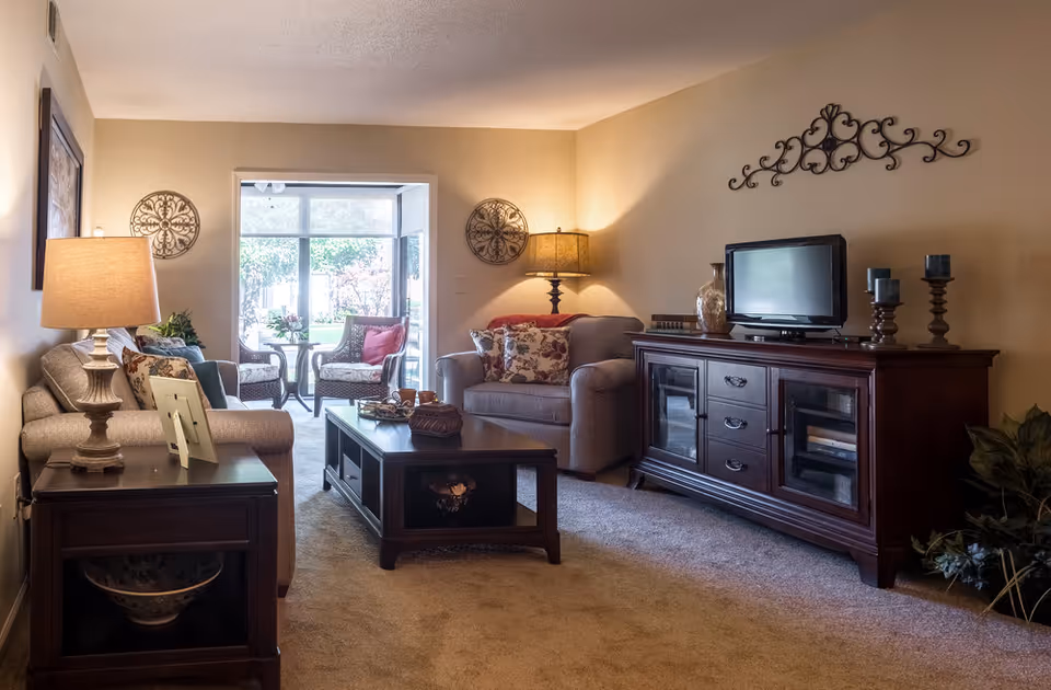 A cozy living room with beige walls and carpet, featuring two upholstered sofas with decorative pillows, a dark wooden coffee table, and a matching TV stand with a flat-screen television. The room is softly lit by two table lamps and decorated with wall art and plants. A sunroom with wicker chairs and a small table is visible through a large windowed doorway.