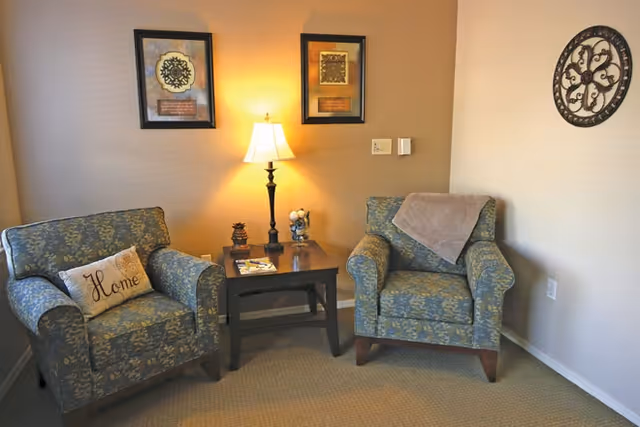 A cozy corner with two patterned armchairs separated by a wooden side table holding a lamp and decorative items. One chair has a pillow with the word 'Home' and the other has a folded blanket draped over the back. The walls are decorated with two framed pictures and a round ornamental wall piece.