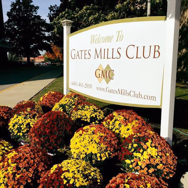A white sign with gold trim that reads 'Welcome To Gates Mills Club GMC' along with a phone number and website, surrounded by colorful yellow, red, and orange flowers. Trees and a sidewalk are visible in the background.