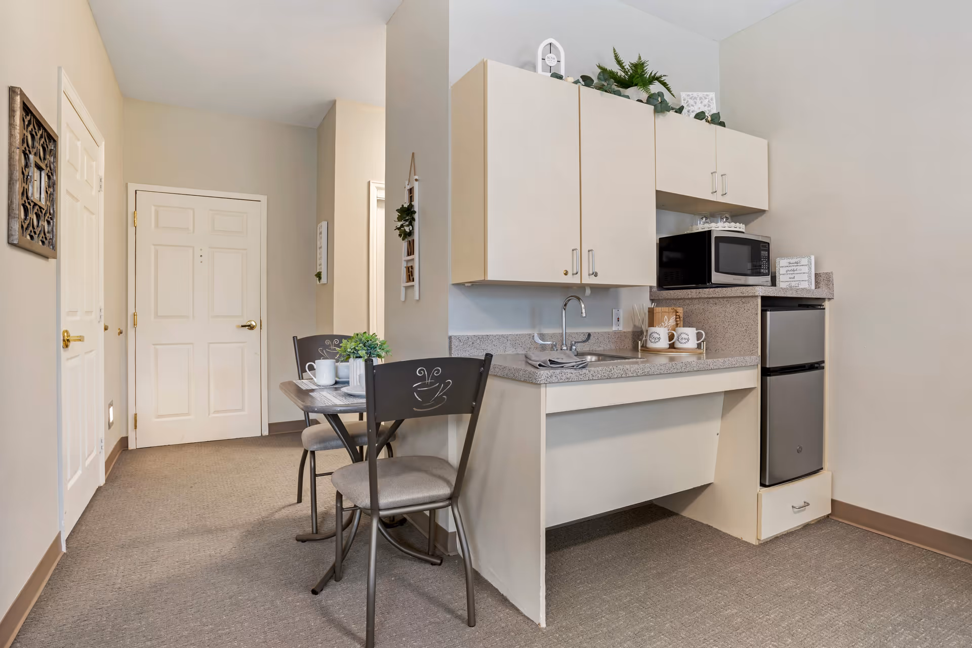 A small kitchenette area with beige cabinets, a countertop with a sink, a microwave, and a mini refrigerator. Next to the kitchenette is a small dining table set with two chairs, each with a coffee cup design on the backrest. The room has beige walls and carpeted flooring, with a door and hallway visible in the background.