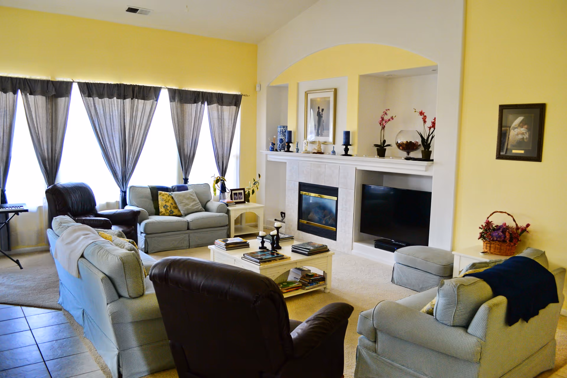 A cozy living room with light yellow walls, multiple sofas and armchairs arranged around a white coffee table filled with books. There is a fireplace with decorative items on the mantel, a flat-screen TV mounted in a recessed wall space, and large windows with sheer dark curtains letting in natural light.