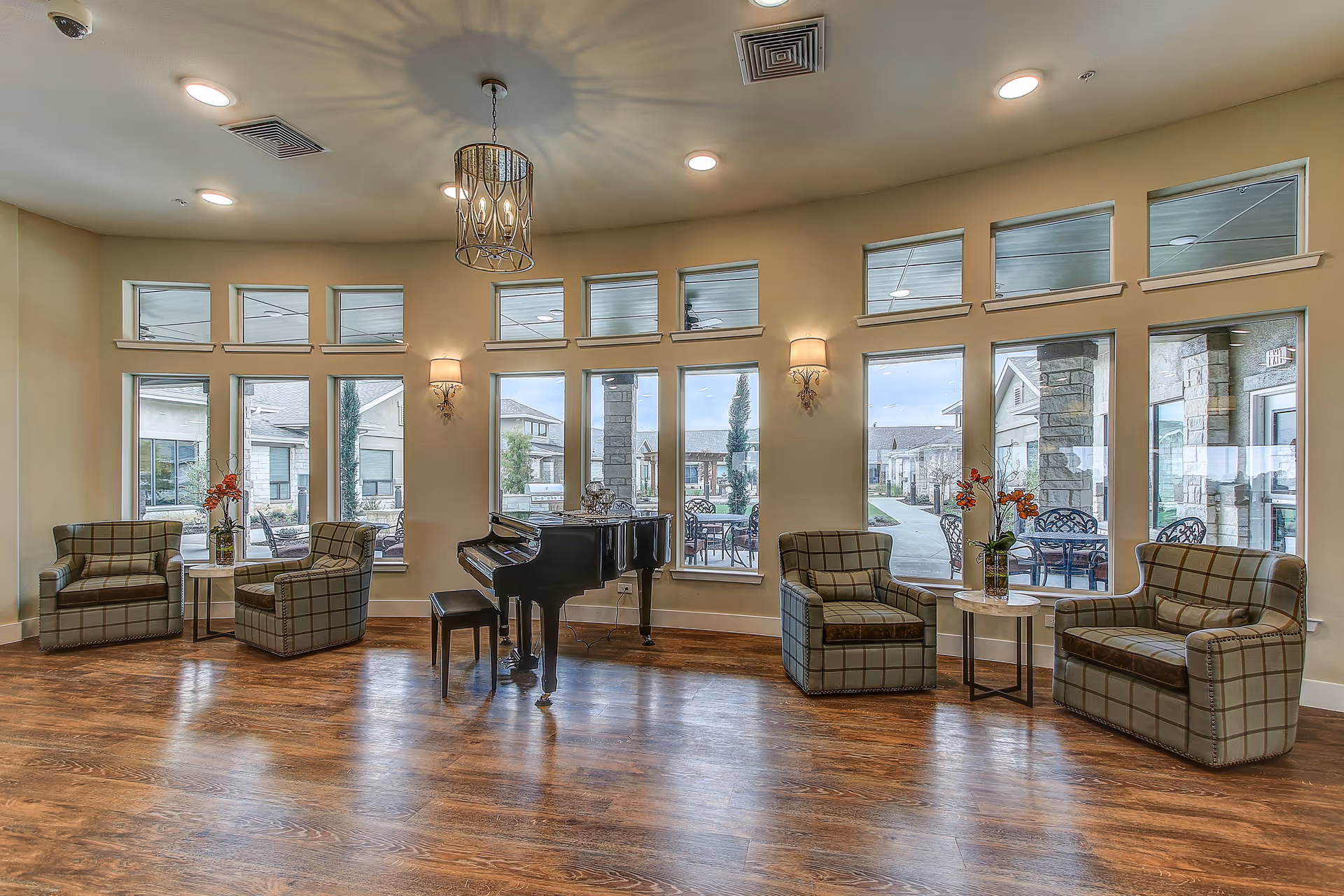 A bright and spacious lounge area with large windows letting in natural light. The room features a black grand piano with a matching bench in the center, surrounded by comfortable plaid armchairs and a loveseat arranged around small round tables with flower vases. The floor is wooden, and the walls are painted a soft beige. Outside the windows, a patio area with tables and chairs is visible.
