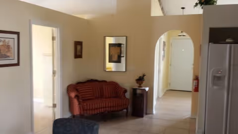 Interior view of a living area with beige walls and tiled floor. A red striped loveseat is placed against the wall, next to a small wooden side table with a decorative item on it. There is an open doorway on the left leading to another room, and an arched hallway leading to a white door in the background. Part of a refrigerator is visible on the right side of the image.