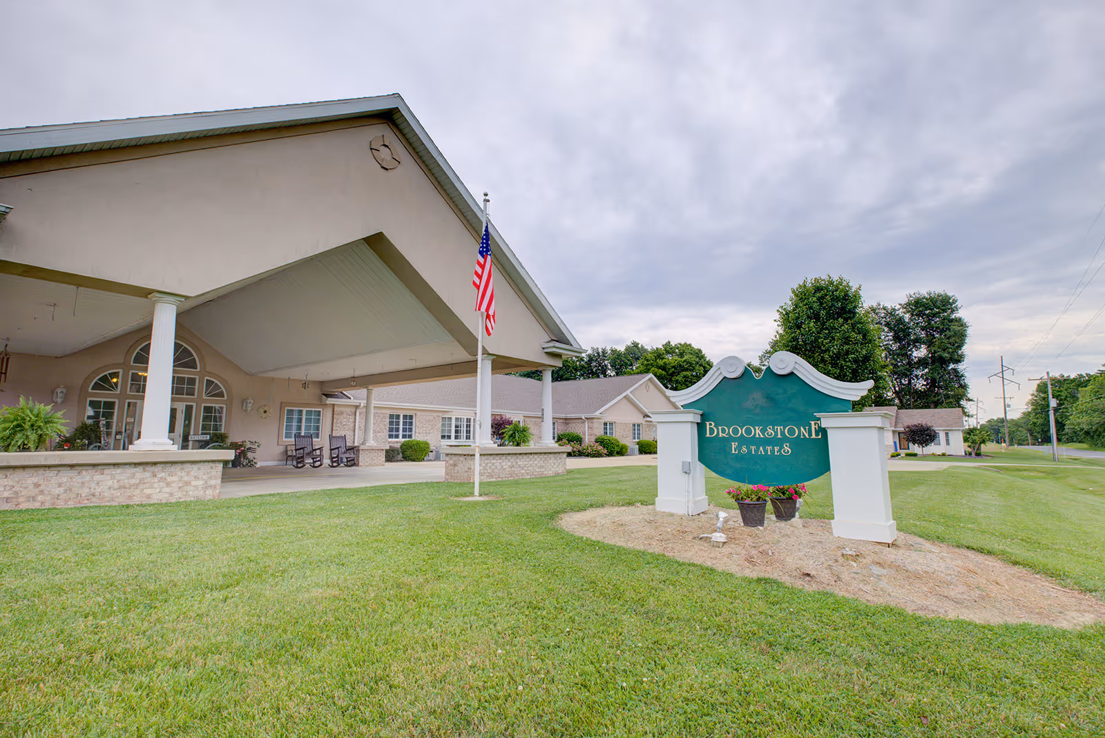 Front entrance of Brookstone Estates showing a covered porte-cochère, American flag, and a green facility sign on a lawn.