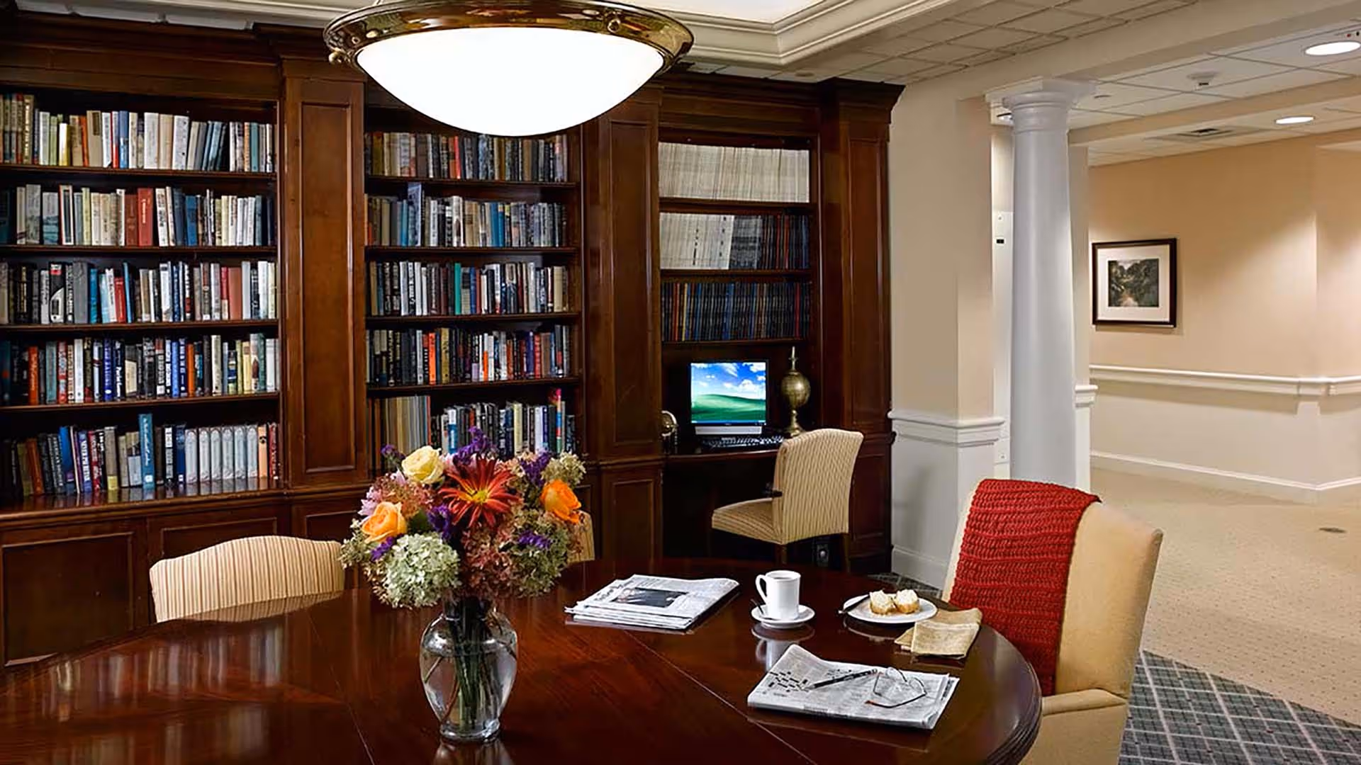 A cozy library-style common room with built-in bookshelves, a large wooden table topped with a vase of flowers, newspapers and cups, and several chairs.