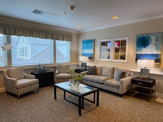 A cozy living room with a beige sofa, two striped armchairs, a glass coffee table with decorative items, two side tables with lamps, and three pieces of wall art. Large windows with blinds and a valance let in natural light.