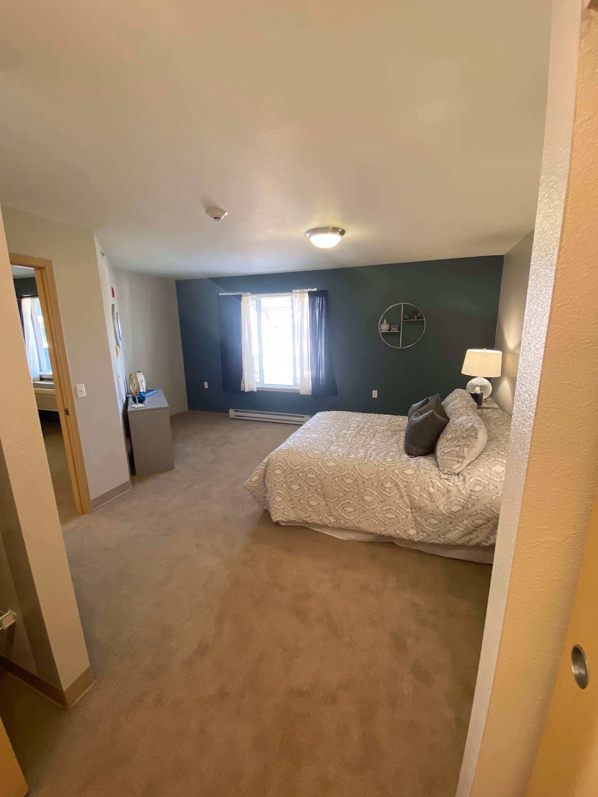 A spacious bedroom with beige carpet and a large bed covered with a patterned white and gray bedspread. There are two pillows on the bed, a bedside table with a lamp, and a round wall shelf on a dark accent wall. A window with curtains lets in natural light, and there is a small dresser near the doorway.