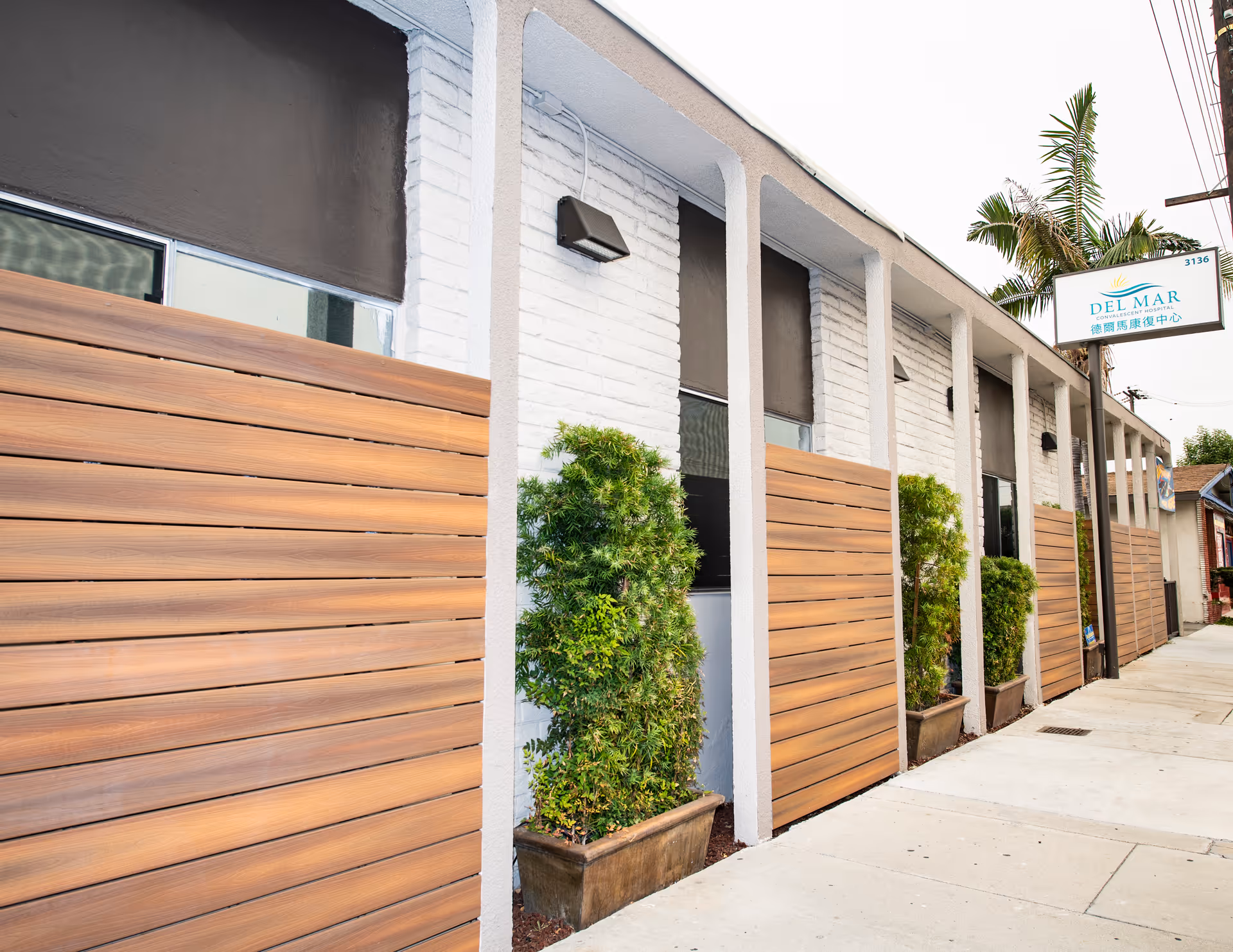 Front exterior of the Del Mar convalescent facility with wood-paneled privacy screens, potted shrubs, and the facility sign above the sidewalk.
