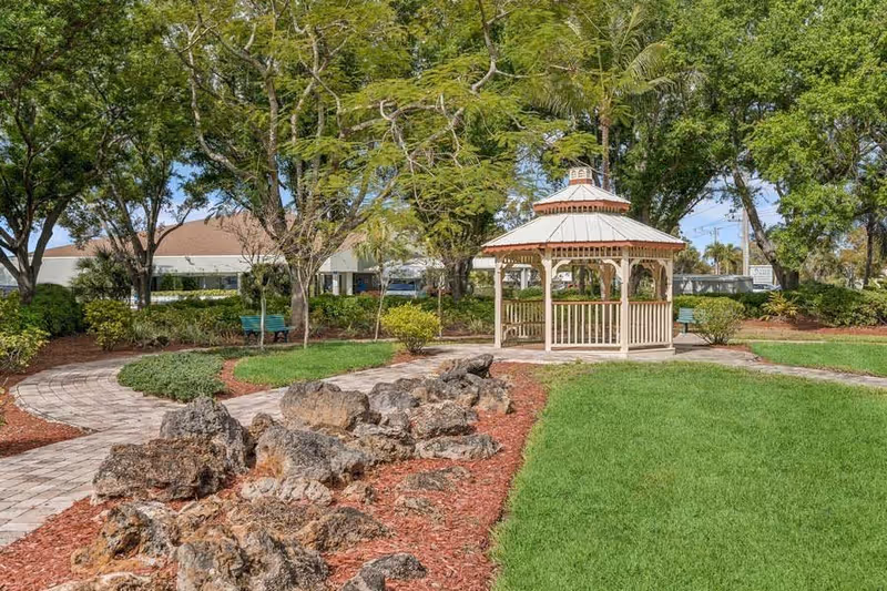 A landscaped outdoor area with a paved walkway, large rocks, green grass, trees, and a white wooden gazebo in the center. There are benches and a building partially visible in the background.