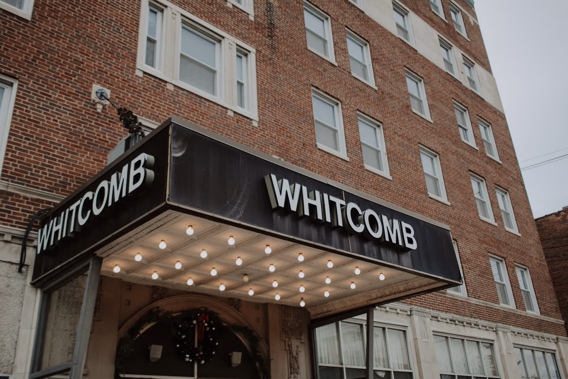 The exterior entrance of The Whitcomb Senior Living Tower, featuring a black marquee with the name 'WHITCOMB' in large white letters and a brick building facade with multiple windows.