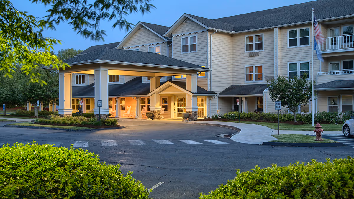 Exterior view of Holiday White Oaks senior living facility at dusk, showing a large covered entrance with warm lighting, beige siding, multiple windows, a flagpole with an American flag, and surrounding greenery.
