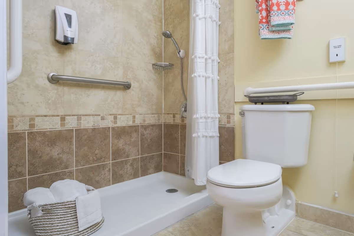 A bathroom with a walk-in shower featuring beige and brown tiled walls, a white shower curtain, a stainless steel grab bar, and a soap dispenser. Next to the shower is a white toilet with a grab bar on the wall behind it. A basket with rolled white towels is placed on the shower floor, and colorful folded towels hang on the wall above the toilet.