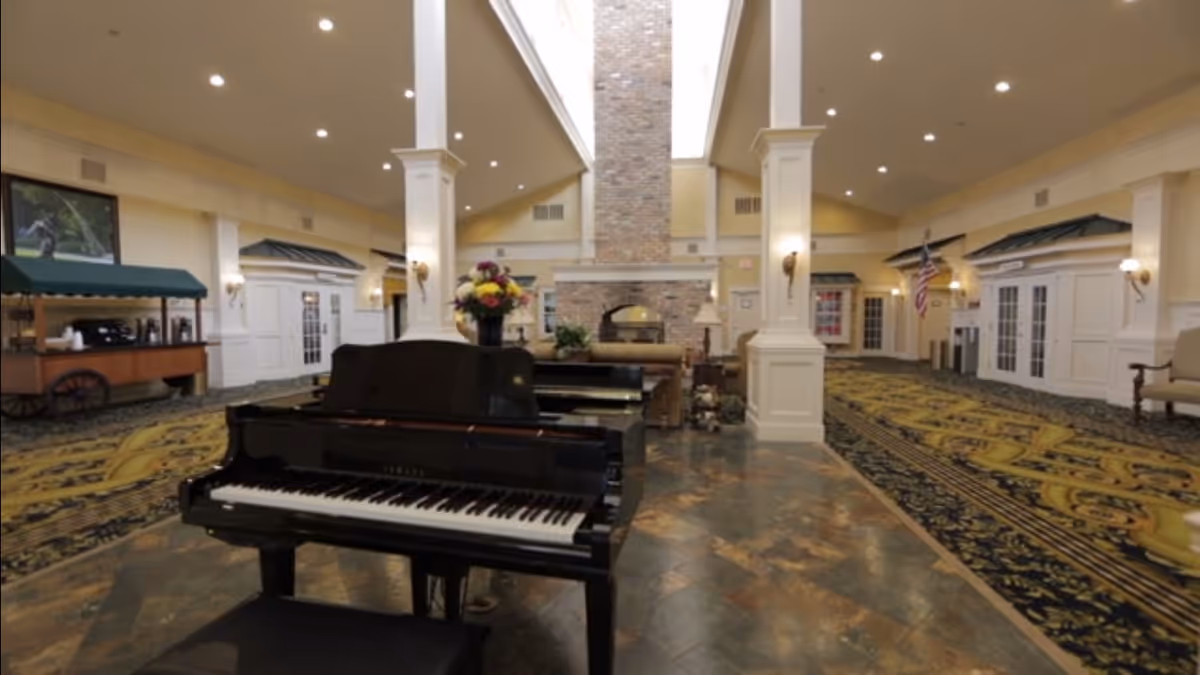 Spacious and elegant common area in a senior living facility featuring a black grand piano with a floral arrangement on top, a large stone fireplace in the background, tall white columns, patterned carpet, and warm lighting.