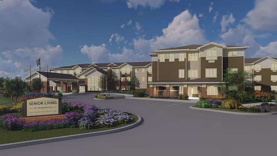 Exterior view of a senior living facility named Senior Living at Towamencin, showing a large multi-story building with a driveway and landscaped garden beds with purple and white flowers in front. The sky is partly cloudy with blue patches.