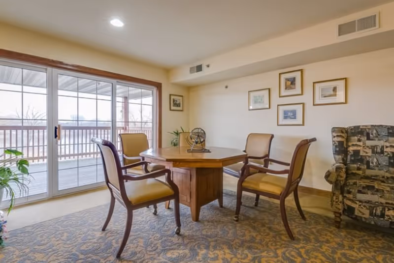 A cozy sitting area with a wooden octagonal table surrounded by four cushioned chairs with wooden arms. The room has a patterned carpet and light-colored walls adorned with five framed pictures. A large sliding glass door leads to an outdoor balcony with a wooden railing. There is also a patterned armchair on the right side and some green plants near the sliding door.