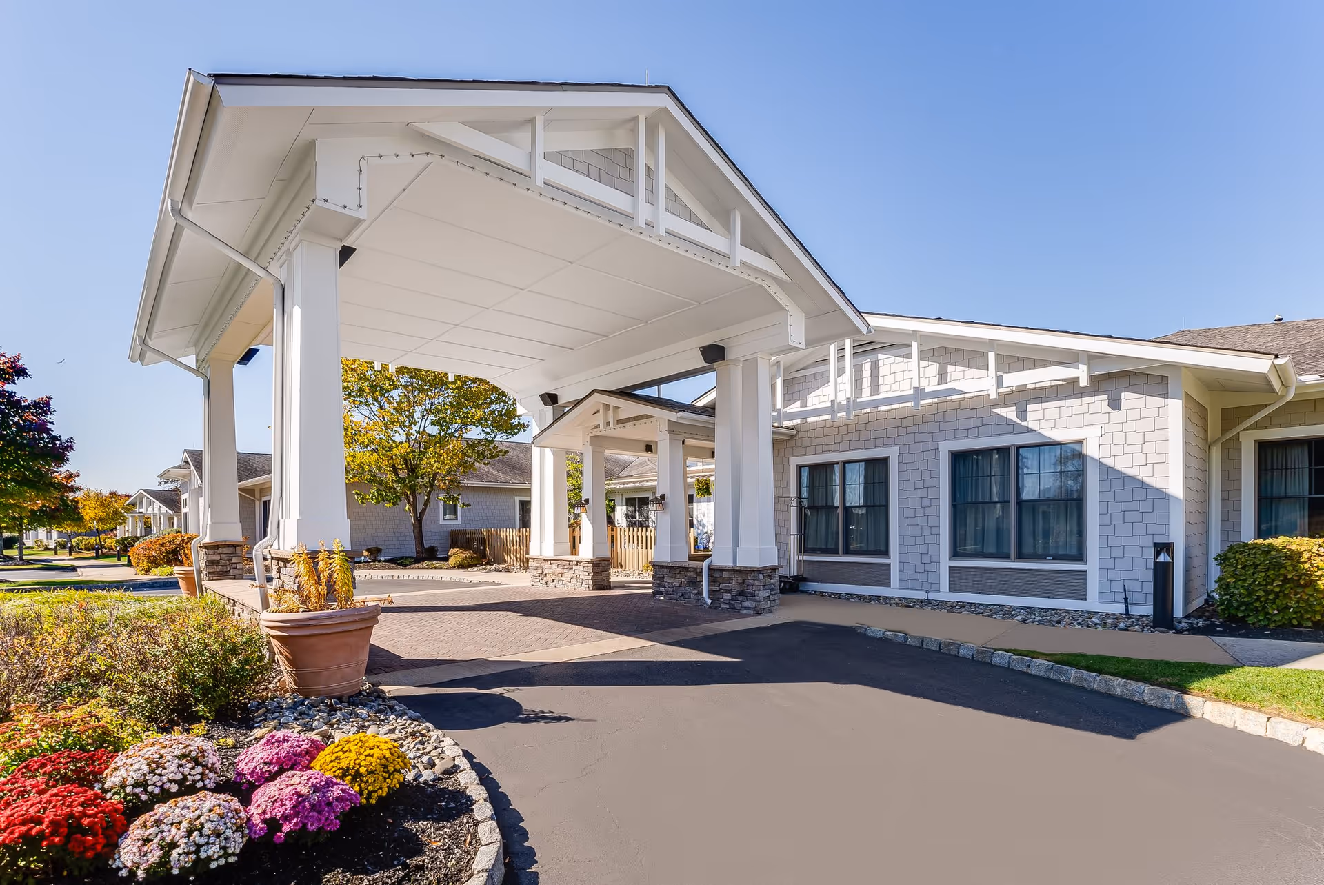Entrance of a senior living facility with a covered drop-off area supported by white columns. The building exterior features light gray siding and multiple windows. There are colorful flowers and landscaping near the driveway under a clear blue sky.