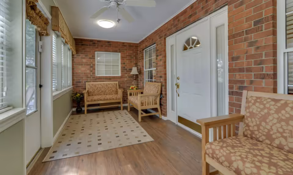 Enclosed bright entryway with exposed brick walls, a white front door, cushioned wooden chairs, a rug, and multiple windows.