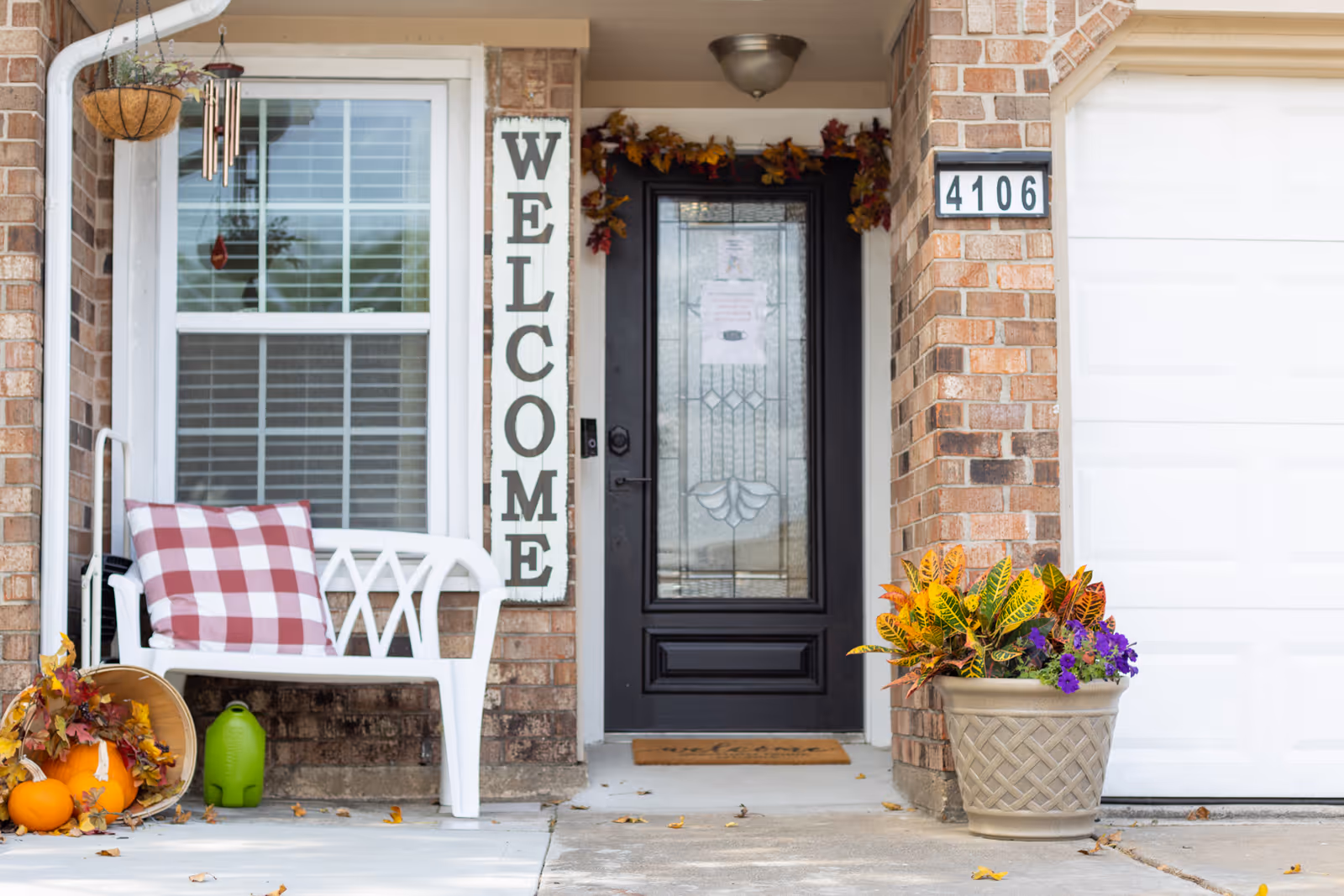 Front entrance of a brick house with a black decorative glass door, a white bench with a red and white checkered pillow, a vertical wooden sign that says 'WELCOME', a hanging basket with plants, a tipped basket with pumpkins and autumn leaves on the ground, and a large planter with colorful flowers next to a white garage door numbered 4106.