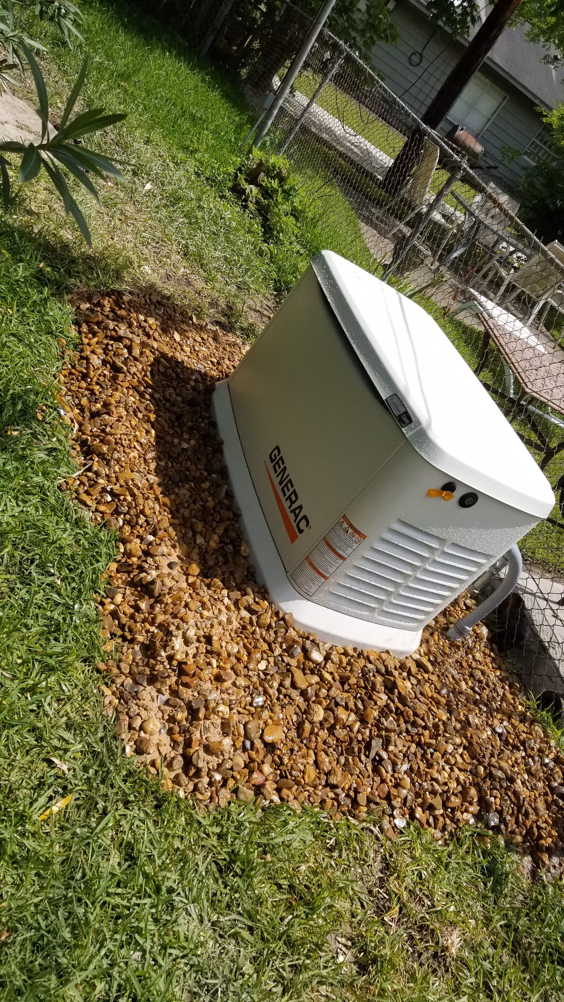 A Generac standby generator installed outdoors on a bed of small brown rocks surrounded by green grass, with a chain-link fence and trees in the background.