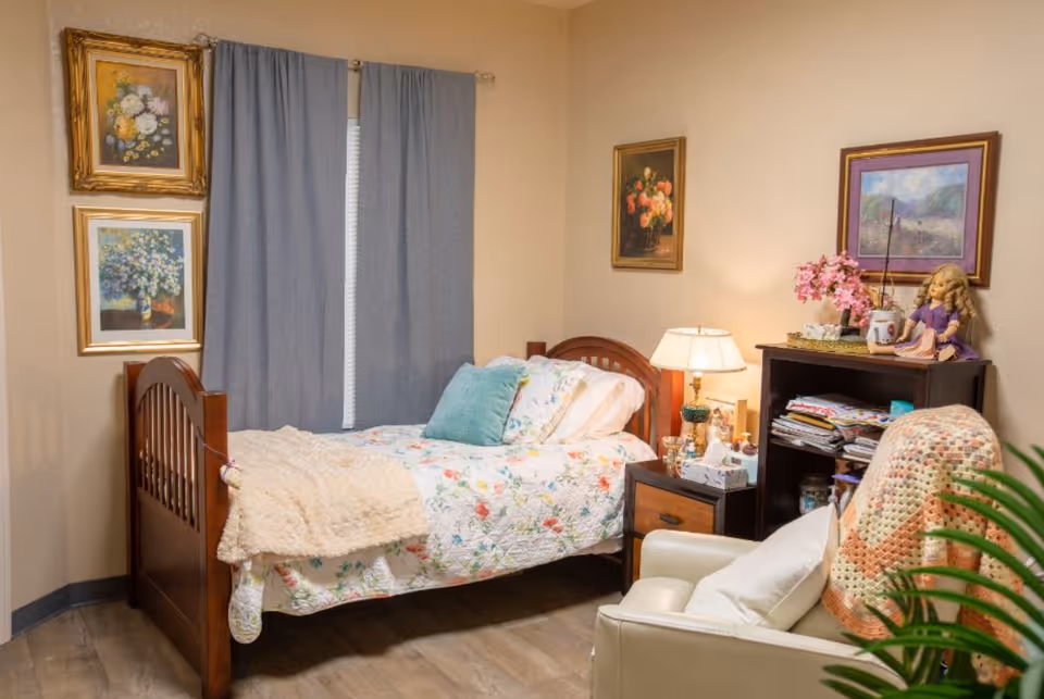 A cozy bedroom with a wooden bed featuring floral bedding and a light blue pillow. Next to the bed is a nightstand with a lamp, tissue box, and decorative items. A comfortable armchair with a crocheted blanket is positioned nearby. The walls are decorated with framed floral paintings, and a window with blue curtains is behind the bed. A small bookshelf holds folded linens and a doll, with a vase of pink flowers on top.
