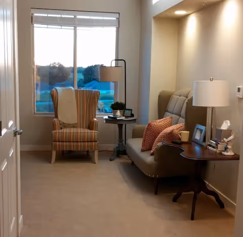 A cozy living room area with a striped armchair near a large window, a small round side table with a lamp and plant, and a cushioned sofa with orange patterned pillows. A wooden side table next to the sofa holds a lamp, framed photos, a tissue box, and a cup.