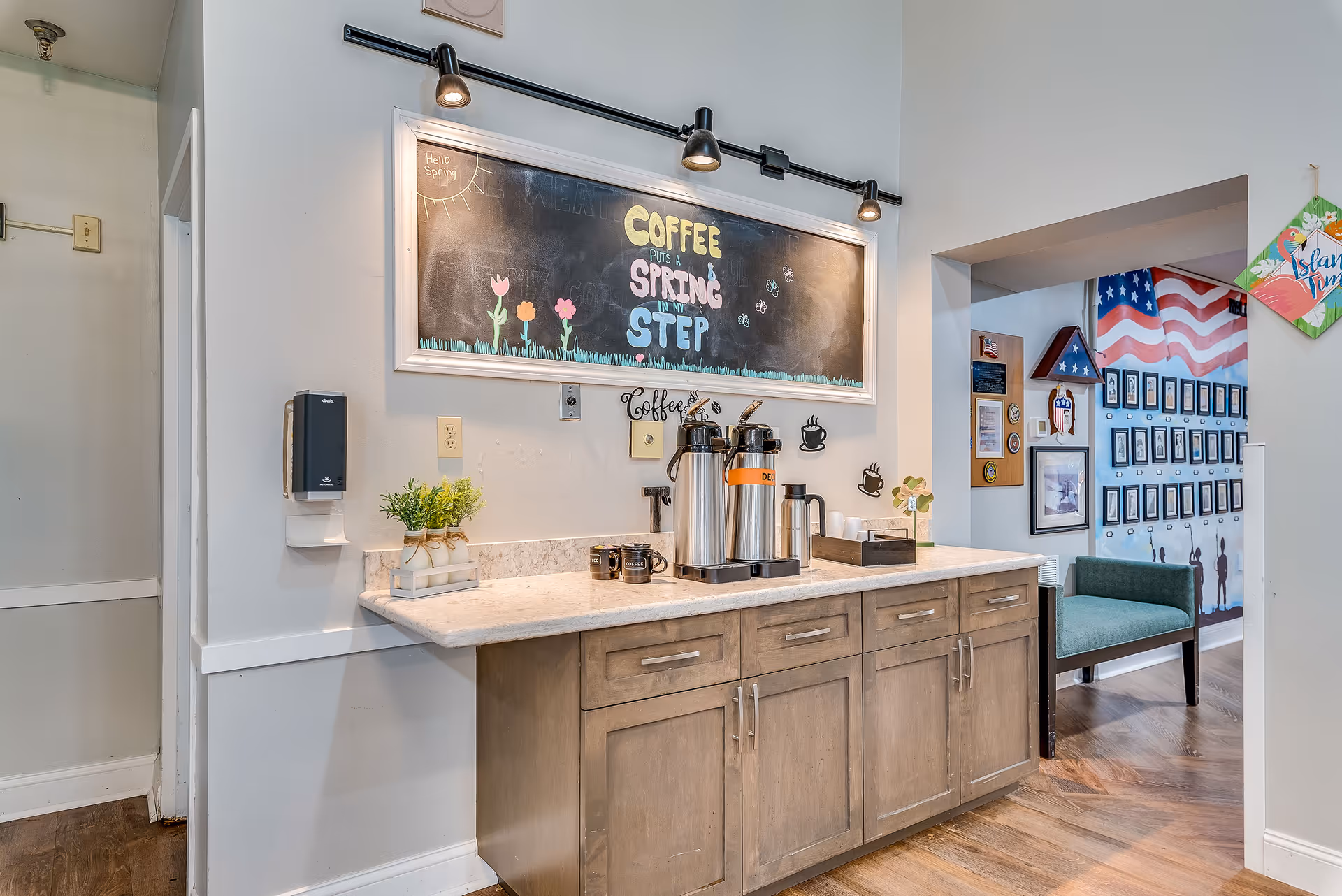 A coffee station in a senior living facility with a countertop holding two large coffee dispensers labeled regular and decaf, coffee mugs, and a small plant. Above the counter is a blackboard with colorful chalk writing that says 'Coffee puts a spring in my step' along with drawings of flowers and butterflies. To the right, there is a hallway with a green bench and a wall decorated with American flags and framed pictures.
