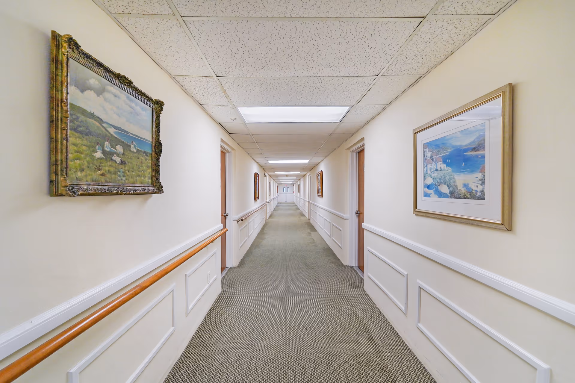 A long, well-lit hallway in an assisted living facility with beige walls, carpeted floor, wooden handrails on both sides, and several framed paintings hanging on the walls. Multiple closed wooden doors line the corridor, and double doors are visible at the far end.