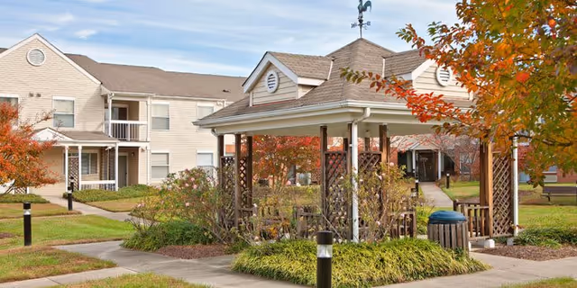 Outdoor view of a senior living facility named Rollingcrest Commons featuring a beige two-story building with balconies, a gazebo surrounded by plants and trees with autumn foliage, paved walkways, and outdoor lighting.
