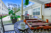 Outdoor covered patio area at The Trace with a round metal table holding a backgammon board, a white coffee cup and saucer, and a small orange candle. The patio overlooks a walkway between two buildings with windows and greenery, including potted plants and hanging ferns. A television is mounted on the wall to the right.