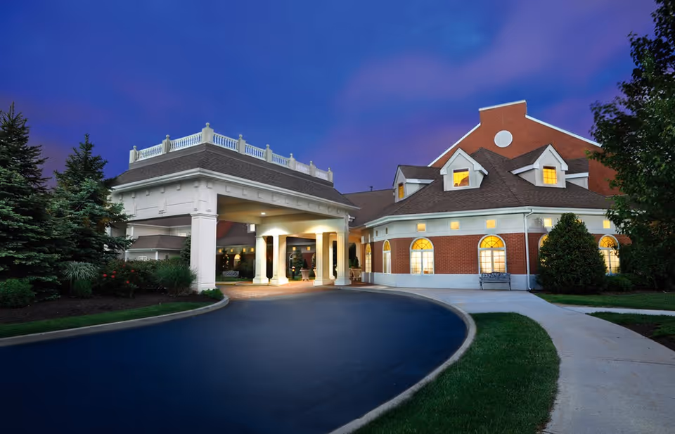 Brick senior living facility front entrance with a covered porte-cochère, curved driveway and landscaped grounds lit at dusk.