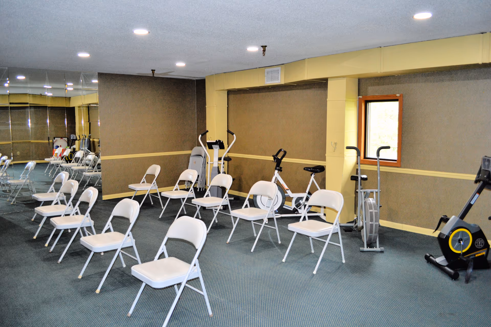 A fitness room with several white folding chairs arranged in rows on a carpeted floor. The room has beige walls with a yellow trim, a small window, and several exercise machines including stationary bikes and a rowing machine. One wall is covered with mirrors reflecting the room.