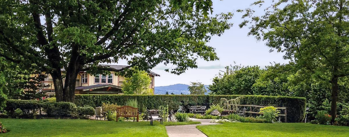 A well-maintained outdoor garden area with green grass, trees, bushes, and a paved walkway leading to a seating area with benches and chairs. In the background, there is a building partially visible behind the trees and hedges, with hills visible in the distance under a clear sky.