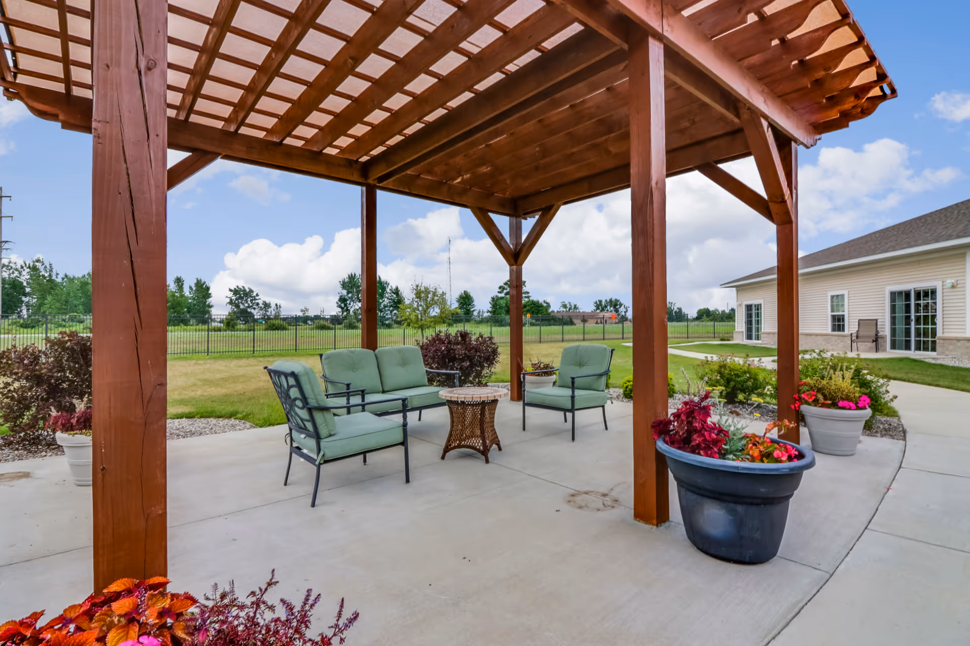 Outdoor seating area under a wooden pergola with green cushioned chairs and a small table, surrounded by potted plants and a well-maintained lawn with a building in the background under a partly cloudy sky.