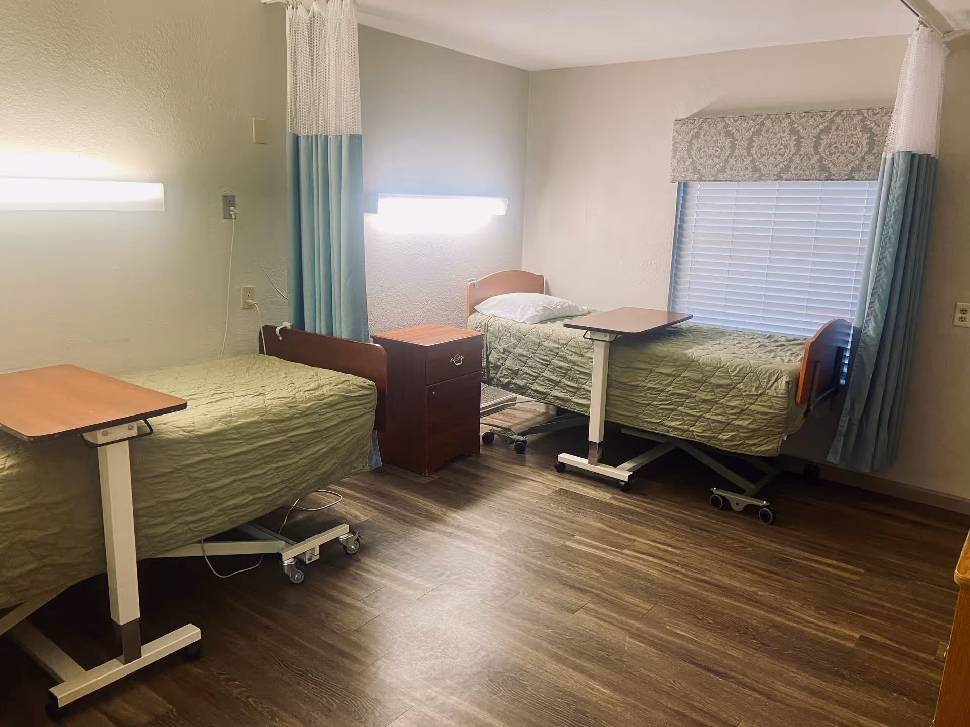 Shared patient room with two hospital-style beds, overbed tables, a nightstand, and a window with blinds.