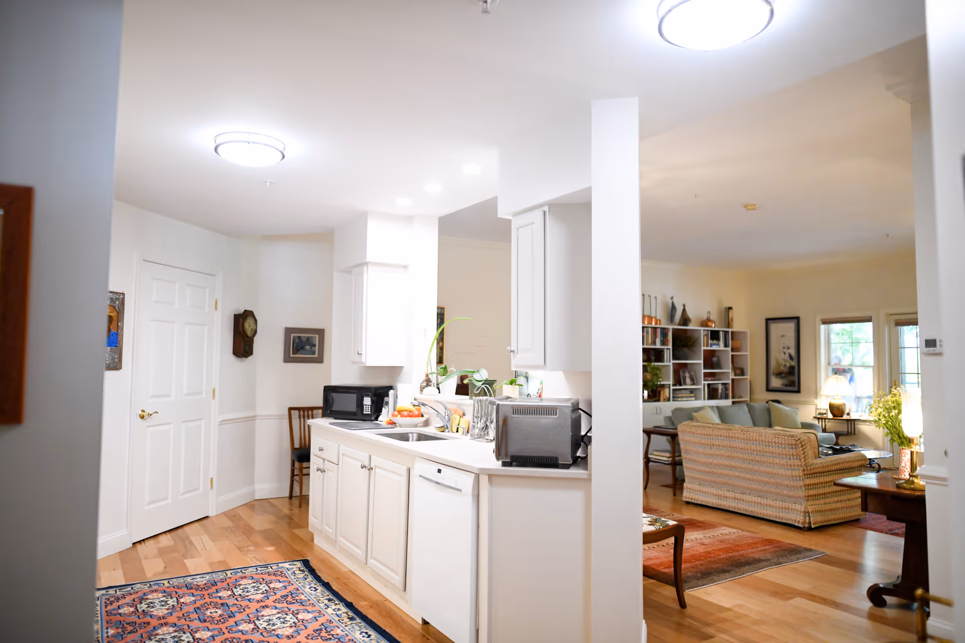 Open-plan interior showing a white kitchen with a sink, microwave and toaster island opening into a living room with sofas and bookshelves.