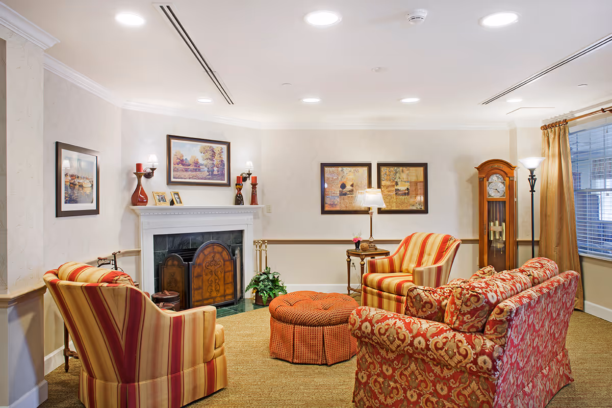 A cozy living room with patterned upholstered armchairs and a sofa arranged around a round orange ottoman. The room features a white fireplace with decorative items on the mantel, framed artwork on the walls, a grandfather clock, and a window with beige curtains. The lighting is provided by ceiling lights and floor lamps.