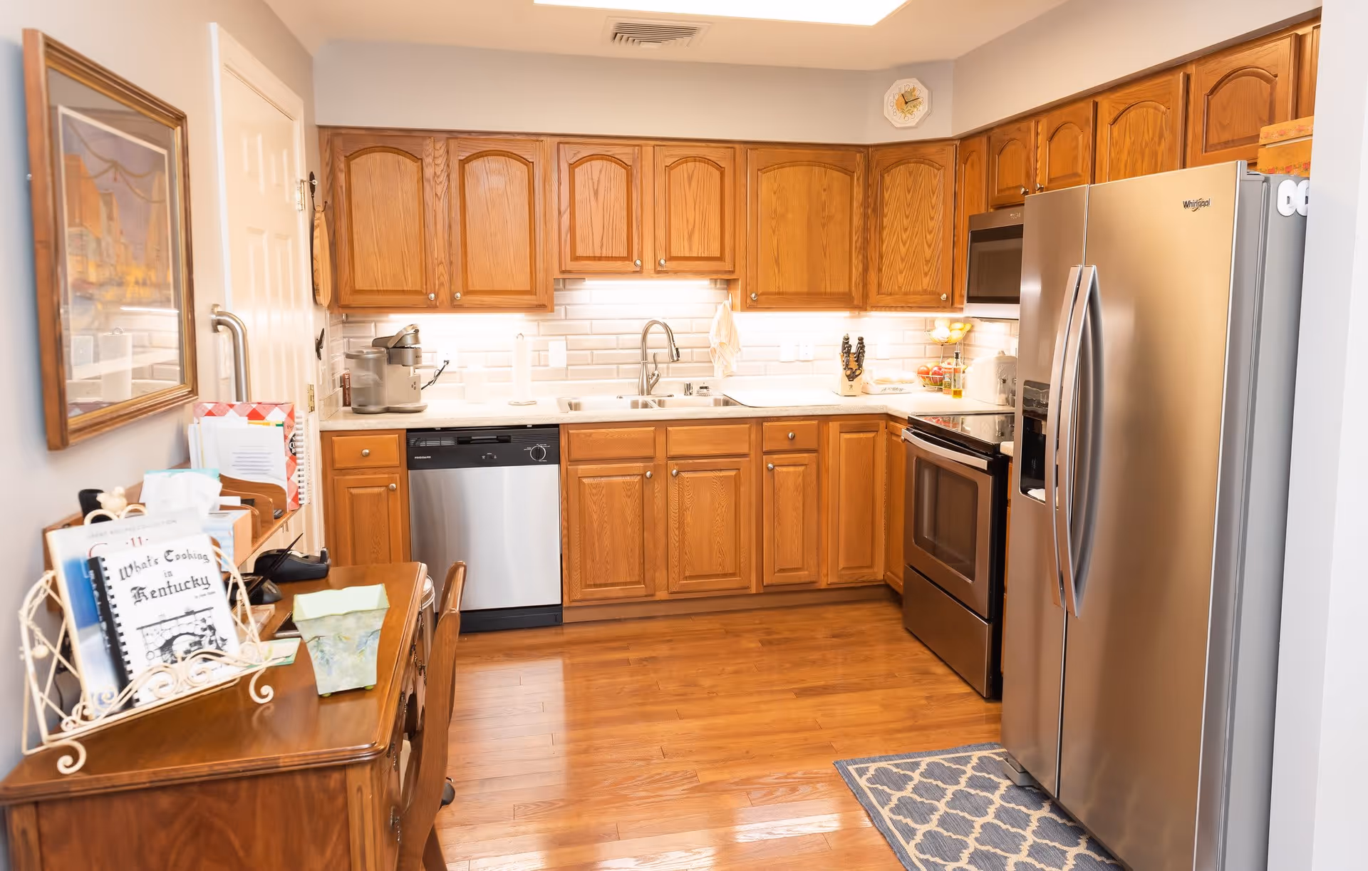 A well-lit kitchen with wooden cabinets, stainless steel appliances including a refrigerator, dishwasher, and oven. There is a double sink under a window, a small rug on the wooden floor, and a small wooden table with books and papers on the left side.