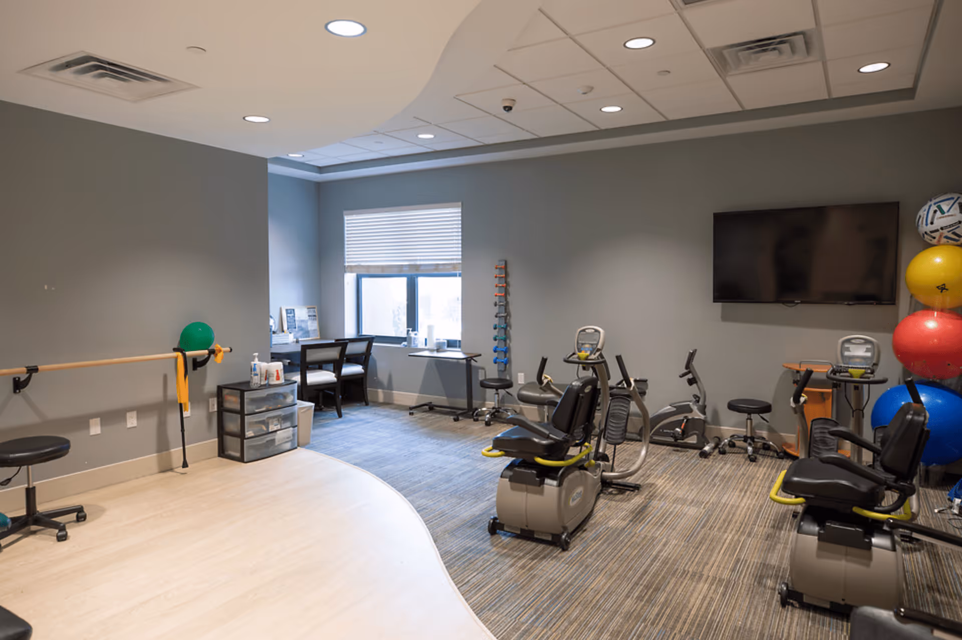 A fitness or rehabilitation room with exercise equipment including stationary recumbent bikes, a ballet barre on the left wall, a rack of colorful exercise balls, a set of small dumbbells, a TV mounted on the wall, and a desk with two chairs near a window with blinds.