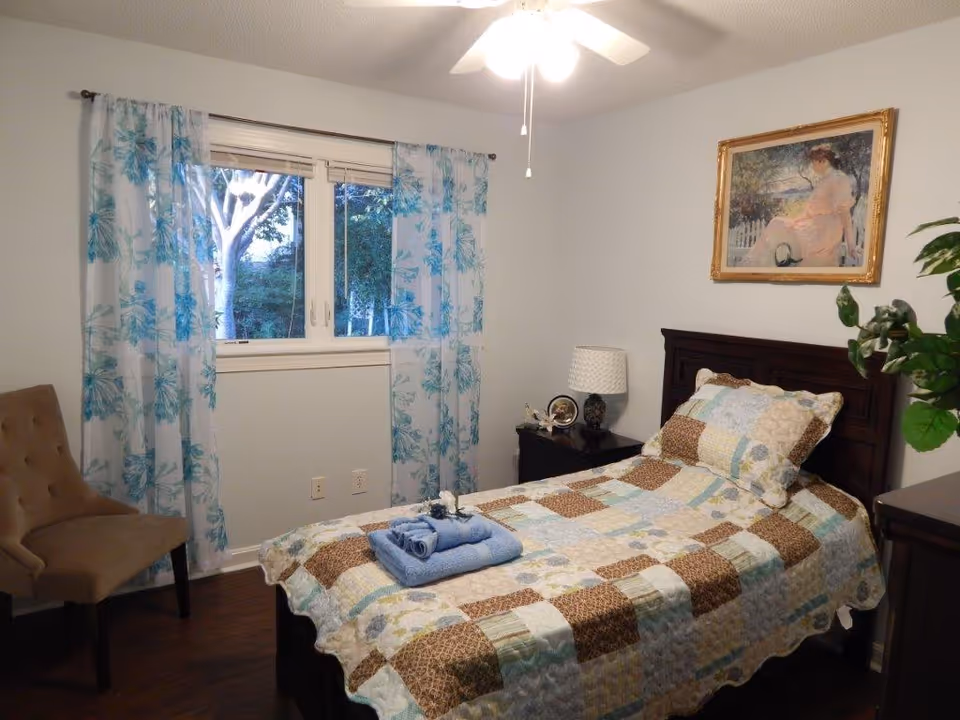 A tidy single-occupancy bedroom with a twin bed covered by a patchwork quilt, a window with blue floral curtains, a chair, nightstand with lamp, and framed wall art.