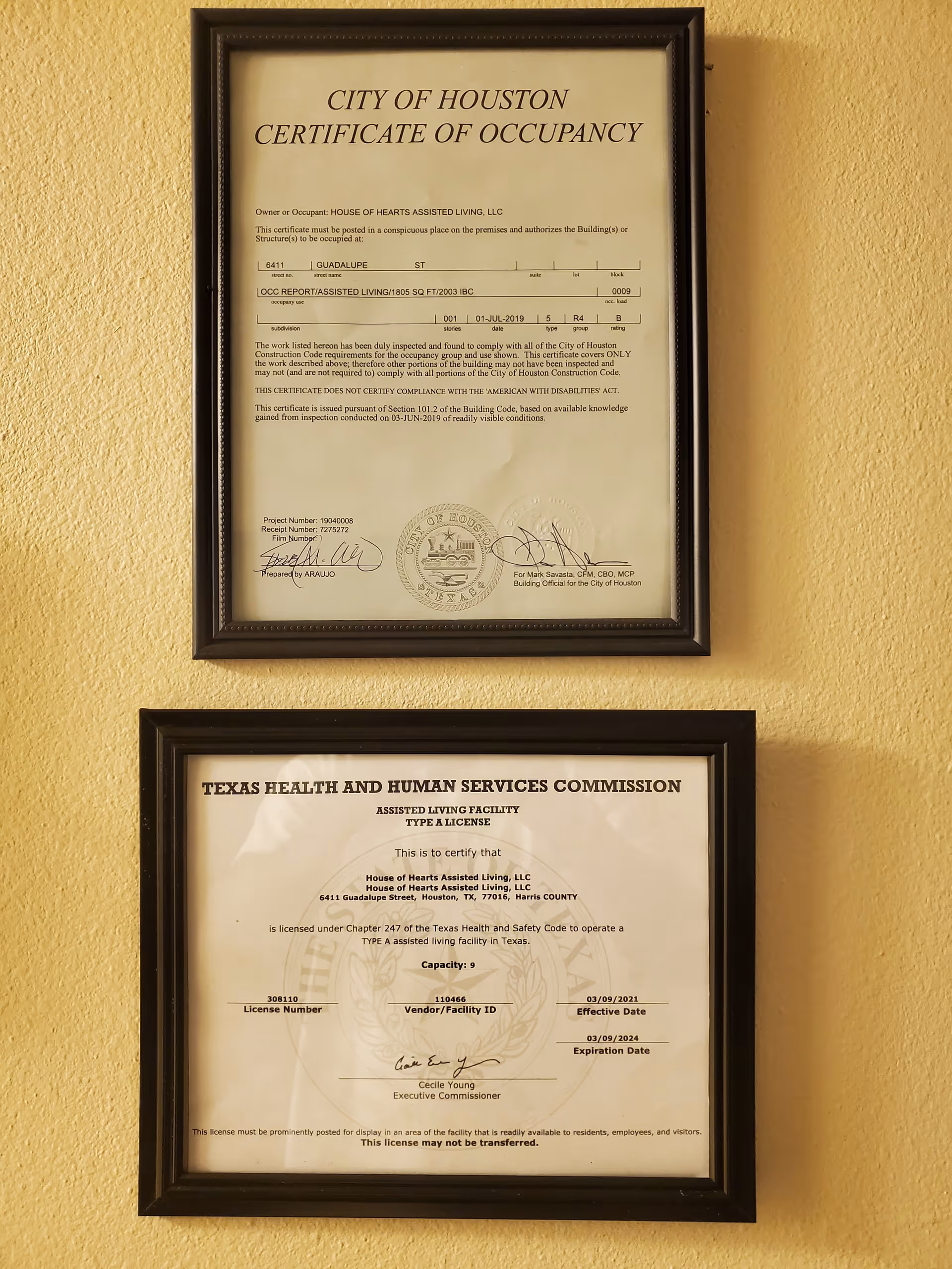 Two framed certificates hanging on a yellow wall. The top certificate is a City of Houston Certificate of Occupancy for House of Hearts Assisted Living, LLC. The bottom certificate is a Texas Health and Human Services Commission Assisted Living Facility Type A License for House of Hearts Assisted Living, LLC.