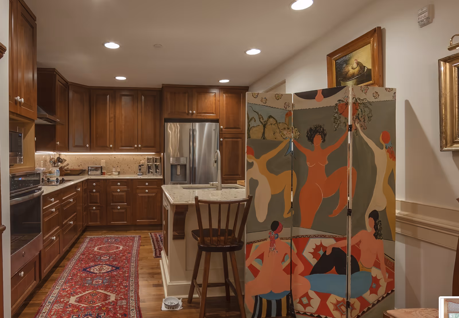 A warm kitchen with dark wood cabinets, a central island and a colorful folding screen painted with figures in the foreground.